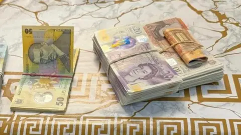 HMRC/Poliția Română Un teanc de bancnote de lire sterline și monedă românească pe o masă